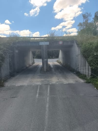 The image shows a concrete overpass with a sign reading "Parc d'Activités de la Croisière" and a red and white circular sign indicating a height limit of 4.3 meters. The overpass is surrounded by vegetation, and a road passes underneath it. The sky is blue with scattered white clouds. The scene appears to be in Saint-Maurice-la-Souterraine, France, as indicated by the presence of the sign. The road leading up to the overpass is paved and has white lane markings. The overall impression is of a rural or semi-rural setting.