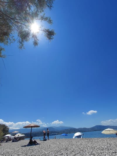 The image depicts a sun-drenched beach with a clear blue sky and a few scattered clouds. In the foreground, a gravelly beach is visible, with several beach umbrellas providing shade. One prominent thatched umbrella is in the center, with a person seated beneath it. Further back, there are other umbrellas, some with people sitting under them. Two men are standing on the beach, one appears to be interacting with a blue inflatable object. In the mid-ground, the calm blue water of the sea stretches towards a distant shoreline with mountains. A large ship is visible on the water in the distance. The sun is high in the sky, casting bright light and some lens flare through the branches of a tree in the upper left corner. The overall scene is one of a typical summer day at a coastal resort. The location is the Municipal Unit of Aegio, Greece.