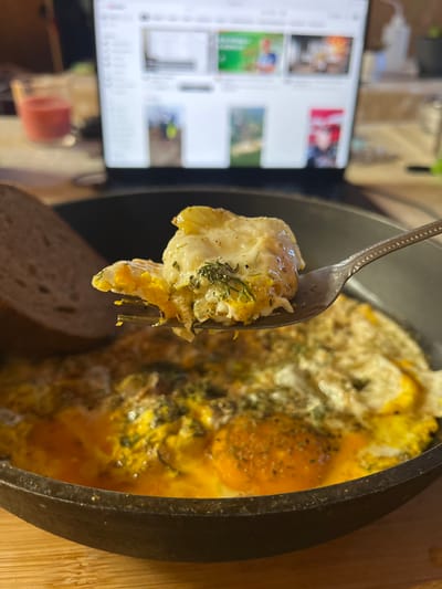 The image depicts a close-up view of a breakfast meal consisting of fried eggs with herbs and a slice of dark bread, served in a cast-iron skillet. A fork is lifting a piece of egg and yolk. In the background, a laptop displays a webpage, possibly a video streaming service, with multiple video thumbnails visible. To the left of the laptop, a glass with a red substance, likely a drink or a candle, is present. The scene is set on a wooden surface. The image appears to be taken indoors. There is no indication of the city of Vasilyevo, Russia, within the provided image.