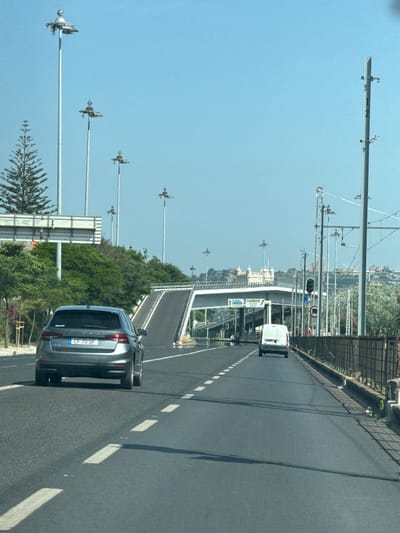 The image shows a view from a vehicle driving on a multi-lane road in Oeiras, Portugal, on a clear day. In the foreground, a silver hatchback car with license plate "CF 70 SF" is visible in the left lane. Further ahead, in the right lane, a white van is driving. The road leads towards an elevated section of road or a bridge, with ramps going up and over. Tall lampposts with multiple light fixtures are spaced along the roadside, and overhead power lines are present. To the left of the road, there are green trees and some vegetation. In the background, a complex of light-colored buildings, possibly a historical landmark, is situated on a hill. The sky is clear and blue.