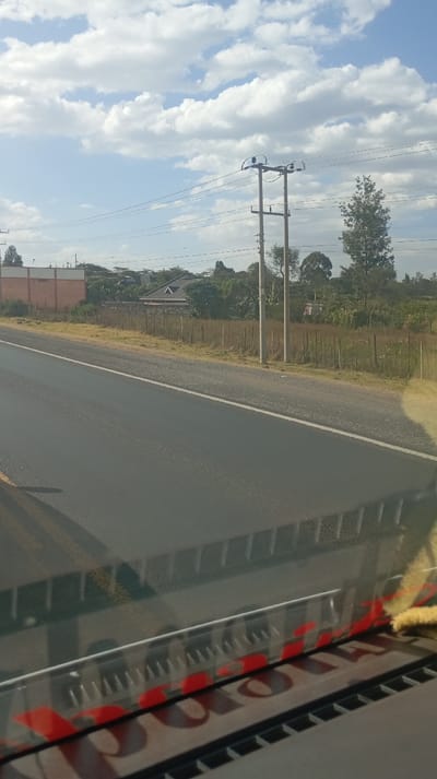 A sunny daytime scene features a black asphalt road with a white dividing line. Adjacent to the road is a grassy area with utility poles carrying power lines. Beyond the grassy area are trees and buildings. The sky is blue with scattered white clouds. The lower portion of the image includes part of the dashboard of a vehicle, including the words "JOURNEY" in red lettering on a clear plastic or glass surface.