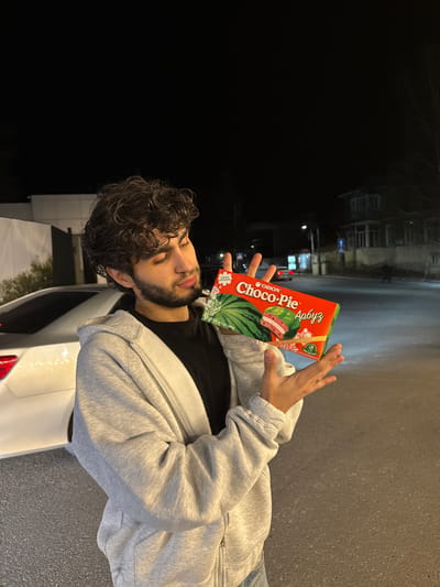 A man with dark, curly hair and a beard is holding a box of Orion Choco-Pie (watermelon flavor) in his hands outdoors at night. The man is wearing a gray sweatshirt over a black shirt and jeans. The location appears to be a street in Tsakhkadzor, Armenia, based on possible identifiable environmental elements in the background. A white car is partially visible. The street and some buildings are faintly visible in the background.