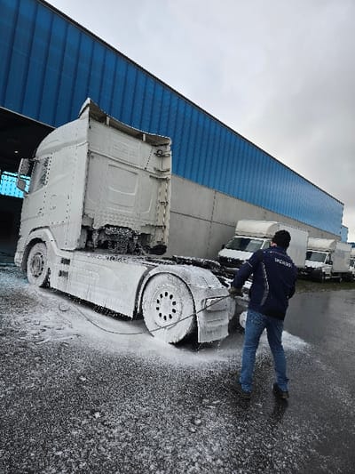 The scene depicts an outdoor commercial or industrial area in Ostend, Belgium, under an overcast sky. A white semi-truck tractor unit is positioned on a dark, wet asphalt surface, heavily coated in white cleaning foam from its cab to its rear axle. A male individual, viewed from the rear, is actively engaged in washing the truck. He wears a dark blue jacket, blue jeans, and a dark beanie, holding a silver-colored pressure washing lance directed towards the truck's rear wheels and chassis. White foam accumulates on the ground around the truck and the individual. In the background, a large building features a prominent blue corrugated metal upper facade above a gray concrete base. To the right of the individual, several white commercial vans or smaller trucks are parked in a line in the middle ground.