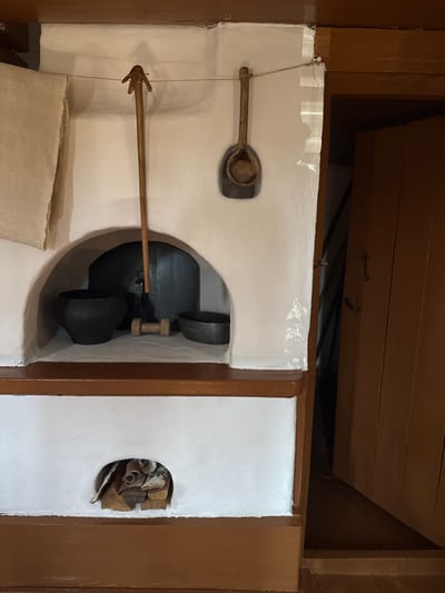 The image depicts a rustic, traditional kitchen scene, likely within a museum or historical reenactment setting in Shushenskoye, Russia. The central focus is a white-plastered stove with a large arched opening. Inside the opening, a large black pot sits prominently, to its left a smaller black pot, and to its right, a shallow metal basin. To the left of the stove, a piece of beige fabric hangs from a string. Above the opening, two wooden tools are suspended: a long-handled tool with a three-pronged head and a smaller wooden spoon. To the right of the stove, a dark wooden door stands ajar, revealing a dark interior. Below the main stove opening, a smaller, circular hearth contains a pile of firewood. The lower portion of the stove and the surrounding frame are painted a dark brown. The overall impression is one of historical domestic life.