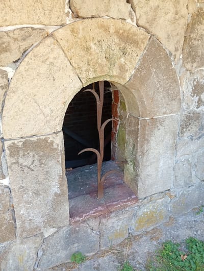 The image shows a close-up view of a small, arched opening in a stone wall. The arch is constructed from rough-hewn, light-colored stone blocks. Inside the opening, a rusty, wrought-iron grate with a branch-like design is visible. The grate appears to be a decorative element rather than a functional security feature. The wall itself is made of irregular, weathered stones of varying sizes and colors, with some patches of green moss and lichen. The ground visible at the bottom of the image is covered with dry grass, small plants, and some gravel. Behind the grate, the interior appears to be dark and made of brick, with some horizontal supports visible. This scene is located at the historical site of the Walkenried Abbey in Walkenried, Germany, likely depicting a detail of its medieval architecture. There are no people or animals present.