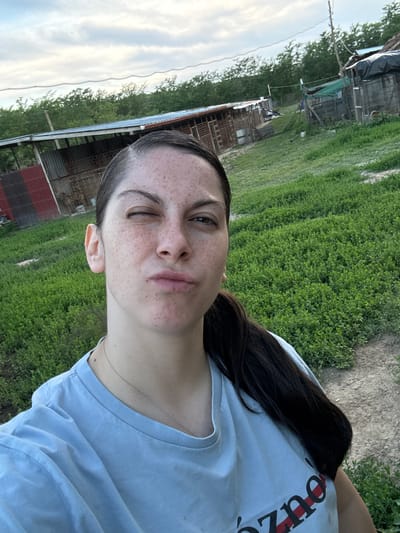 A young woman with brown hair, freckles, and fair skin is taking a selfie outdoors. She is wearing a light blue t-shirt and a thin necklace. She is winking with her left eye. The background consists of a green grassy area, a dirt path, and several dilapidated wooden buildings, possibly sheds or barns, with parts covered in dark tarps or plastic. The sky is visible in the upper left corner and is light with some clouds. The location appears to be rural or a neglected area. The city of Dekov, Bulgaria, is not discernible from the image.