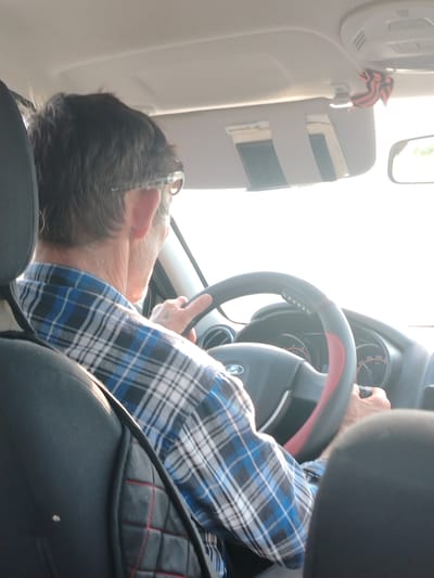 A man is driving a car. He is wearing a blue and white plaid shirt and glasses. The car's interior is visible, including the steering wheel and dashboard. The sun is shining brightly, creating a bright glare. The location is the city of Novyi Shagirt, Russia.