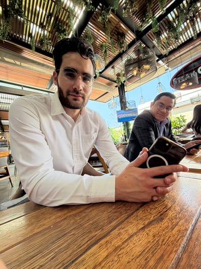 The image displays two males and a partially visible third individual seated at a wooden table in an outdoor or semi-outdoor restaurant patio during clear daytime conditions. Overhead, a wooden slat canopy is adorned with hanging artificial greenery and features several modern pendant light fixtures.

In the foreground, a male with dark hair, a beard, and round, light-rimmed glasses, wearing a white long-sleeved button-down shirt, looks directly at the camera with a neutral expression. His hands are visible in the lower center, holding a dark-colored smartphone horizontally. The phone's rear case exhibits a distinct white circular magnetic accessory.

To the right and slightly behind the foreground subject, a second male wearing glasses and a dark patterned jacket is seated, looking towards the left or downwards while interacting with his own smartphone. A third person's head and shoulder are partially visible on the far right edge of the frame.

The background includes a large flat-screen display exhibiting a landscape image and a partial black sign with white lettering, possibly reading "STEAK CO." Distant green foliage is also visible. The specific location within Santiago de Querétaro, Mexico, cannot be determined from the visual information provided.
