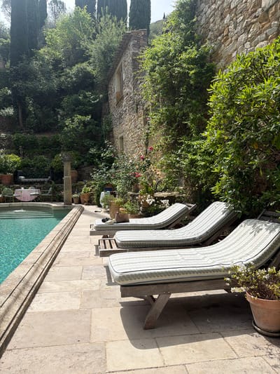 This media file presents a sun-drenched outdoor scene depicting a luxurious poolside area in Saint-Victor-la-Coste, France. The primary focus is a clear, turquoise swimming pool with a light-colored stone-paved patio surrounding it. Along the pool's edge, three wooden sun loungers with striped white and light grey/green cushions are neatly arranged, casting distinct, long shadows that indicate it is likely mid-morning or early afternoon under bright, clear skies.

The setting is characterized by abundant natural beauty and traditional architecture. A large stone building, possibly a historic residence, stands in the background on the left, its walls partially obscured by dense, vibrant green foliage and climbing plants. Various trees, including tall, slender cypress trees on the far left, contribute to the lush, verdant environment. Stone walls, heavily integrated with greenery, define the property's boundaries.

Further into the scene, a smaller poolside seating area is visible, featuring a table draped with a pink tablecloth and two chairs, suggesting a tranquil spot for dining or relaxation. A decorative stone column stands nearby. Potted plants, including one terracotta pot with white flowering blooms beside the loungers, add touches of floral charm. The overall impression is one of serene luxury and a peaceful retreat, showcasing the beautiful natural and architectural elements typical of the French countryside. No people or visible text are present in the image.