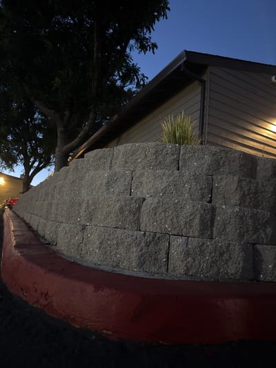 The image depicts a retaining wall made of grey concrete blocks, constructed in a stepped pattern. The wall is bordered at its base by a red-painted curb, which shows signs of chipping and wear. Behind the retaining wall, a building with horizontal siding and a dark brown roof is visible. A single light fixture illuminates a portion of the building's exterior. Above the building, a large tree with dark foliage is silhouetted against a dark blue twilight sky. The scene appears to be an outdoor residential or commercial area, possibly in the city of Sacramento, United States, as indicated by the context. The lighting suggests the image was taken shortly after sunset or before sunrise.