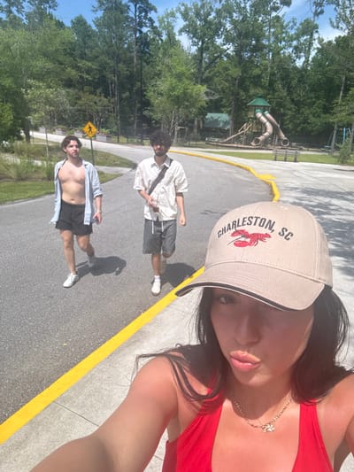 The image shows a woman taking a selfie in a park in Goose Creek, South Carolina, at coordinates 33.053, -79.953. She is wearing a red top, a gold necklace, and a baseball cap with "Charleston, SC" and a lobster design embroidered on the front. Her long dark hair is visible, and she is looking into the camera. Two men are walking in the background on a paved path with a yellow painted line on the edge. One man is wearing a blue shirt with the sleeves rolled up, dark blue shorts, and white sneakers. The second man is wearing a white button-down shirt, black and white striped shorts, and white sneakers. Behind the men is a grassy area, a playground, and a dense forested area.