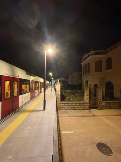 The image displays a nighttime scene at the Picanya metro station platform in Picanya, Spain. A white and red train, marked with "GENERALITAT VALENCIANA" and "Deixeu eixir / Dejen salir" text, is stopped on the tracks, parallel to a grey tiled platform featuring a yellow tactile safety strip. The train's interior lights are active, visible through its windows. A prominent streetlight stands on the platform, centrally located, emitting significant lens flare across the dark sky. Beyond the platform, a low stone wall with decorative pilasters and a black metal fence separates the station area from a narrow, unpaved strip of ground. Directly across from the platform and unpaved area stands a two-story residential building with light-colored stucco, featuring a distinct rounded corner, arched window frames, and some obscured windows, likely by shutters. Further down the street, additional buildings are partially visible in the low light. A circular manhole cover is present on the unpaved ground in the lower right quadrant. No individuals or discernible activities are depicted in the scene.