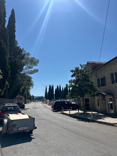 A bright, sunny day with a clear blue sky over a street in Lepetane, Montenegro. Tall cypress trees line the left side of the road, casting shadows on a stone wall. A grey trailer attached to a grey car is parked on the left side of the road, its license plate reading "TV AU 891". On the right side of the road, a stone building with green shutters is visible, with a sign above the entrance reading "AMPHORA". Cars are parked along the right side of the street and driving down the road in the distance. The street is paved and appears to be in good condition. The sun is shining brightly, creating lens flares in the upper part of the image.