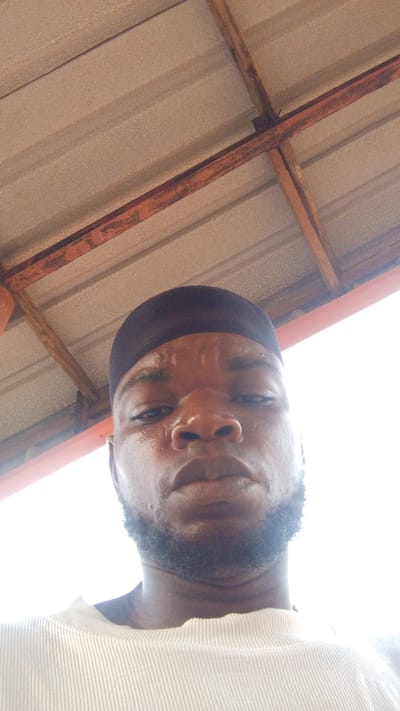 The image is a low-angle selfie of a man with dark skin, a closely cropped beard, and a black head covering. He appears to be perspiring. He is wearing a white, ribbed shirt. Above him is a roof constructed of corrugated metal panels supported by rusty metal beams. The photograph was taken in an outdoor setting with bright, diffused light. The location is speculated to be the city of Enugwu-Ukwu, Nigeria.