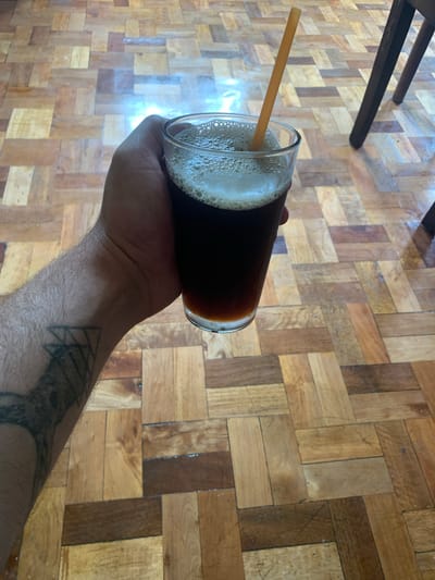 A hand is holding a glass of dark liquid with a straw, resting on a parquet floor. The hand has a tattoo on the forearm and wrist. The glass contains a dark beverage with foam on top, and a straw is inserted into it. The floor is made of wood planks arranged in a parquet pattern, with varying shades of brown and tan. In the background, the legs of a dark wooden chair are visible. The scene is indoors. There is no information in the image to suggest the location is in Chiatura, Georgia.