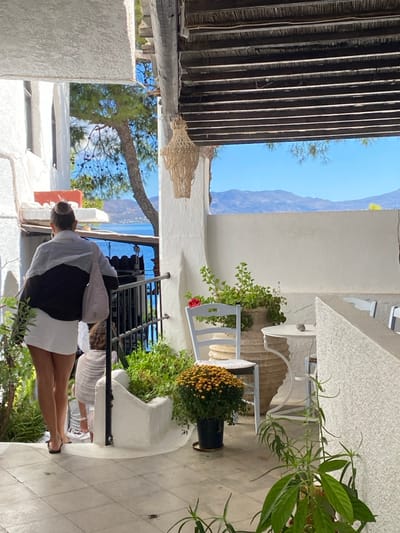 The image depicts an outdoor patio or walkway in Megalochori, Greece, featuring traditional white-painted architecture and a distant sea view. In the left midground, an adult female is seen from behind, wearing a bi-colored long-sleeved top (dark lower, light upper), white shorts, and dark flip-flops. Her hair is styled in a bun, and she carries a light-colored tote bag on her left shoulder. She is descending a short flight of white steps with a black metal railing. A small child, mostly obscured by the adult, follows closely behind her on the steps, wearing a light-colored garment.

To the right, a dedicated seating area is visible with whitewashed walls. It includes a light blue wooden chair with a round white cushion, and a small white round metal table with a dark grey pebble or rock resting on its surface. Numerous potted plants decorate the area, including a prominent pot of orange chrysanthemums, a large terracotta-style pot containing green leafy plants and a single red bloom, and various smaller green plants in white built-in planters.

Overhead, a rustic wooden beam and slat roof structure is visible, from which a tiered, light-colored macrame chandelier hangs. Beyond the railing on the left, a large pine tree partially frames the background vista, revealing a calm blue sea and distant undulating blue mountains under a clear azure sky. The scene is illuminated by bright daylight, suggesting a sunny day.