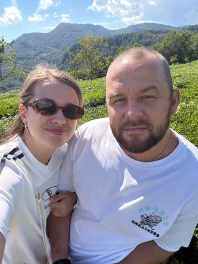 In the city of Izmaylovka, Russia, a man and a woman are captured in a selfie taken outdoors. The man, who appears to be in his late 30s or early 40s, has a shaved head, a beard, and is wearing a white t-shirt with a dragon graphic and Japanese characters that read "GREATNESS." The woman, younger and with light brown hair, is wearing sunglasses and a white t-shirt with black stripes on the sleeve. She is wearing a silver hoop earring and has a purse strap visible. The background reveals a lush, green mountainous landscape under a bright blue sky with scattered clouds. The terrain is covered in dense vegetation, suggesting a natural, possibly rural or mountainous area.