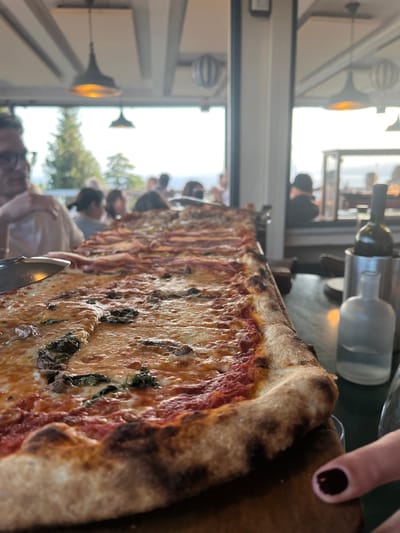 The image captures a long, rectangular pizza, indicative of "pizza al metro" or "pizza alla pala" style, served on a dark wooden board in what is identified as Focacceria e Pizzeria L'Orizzonte in Genoa, Italy. The pizza dominates the foreground and extends into the midground, displaying various toppings including a section with tomato sauce, melted mozzarella, and fresh basil leaves, and further back, sections with cured meats, possibly prosciutto, and other toppings. The crust is visibly well-baked with characteristic charring.
In the blurred background, through large, clear windows, an outdoor panoramic view is visible, featuring lush green trees against a bright, hazy sky, consistent with an elevated dining location overlooking the city or coastline. Indistinct figures of multiple diners occupy tables in the background, appearing to be conversing and enjoying the setting. A person's blurred profile, wearing glasses, is visible on the far left. On the table to the right of the pizza, a dark green wine bottle is partially submerged in a silver ice bucket, accompanied by a clear, frosted glass bottle. On the bottom right, a finger with dark nail polish is visible, pointing towards the pizza. Overhead, modern, bell-shaped pendant lights with warm glowing interiors are suspended from a light-colored, paneled ceiling.