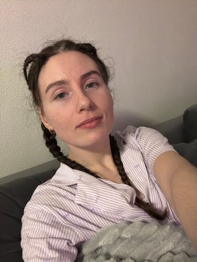 A female individual is depicted in an indoor setting, located in Yanino-1, Russia. She is seated or reclined against a dark gray sofa, with a light-colored, textured wall behind her. Her dark brown hair, exhibiting visible gray roots, is styled into two braided pigtails, which originate from two small buns or knots positioned on the top of her head, with the braids extending downwards. She is wearing a light purple and white striped long-sleeved collared shirt, detailed with small purple bow motifs. A small golden hoop earring is visible in her left ear, and a thin silver chain necklace is present around her neck. Her facial expression is neutral, and she maintains direct eye contact with the camera. A gray, plush blanket covers her lower torso and left arm.