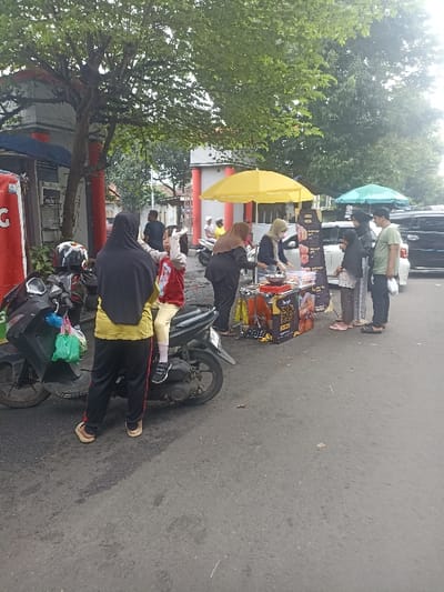 The scene takes place in a street in Purwokerto, Indonesia, at the coordinates -7.417 latitude, 109.249 longitude. There is a street food stall with a yellow umbrella and black sign. Four women are standing at the stall, one of whom is wearing a hijab and another is wearing a yellow shirt. There is another woman standing in the background. A young girl is wearing a red shirt and sitting on a motorcycle. Three men stand to the right of the stall, one of whom is wearing a green shirt. A black car can be seen in the background. The ground is asphalt. The scene is outdoors. The weather appears overcast.