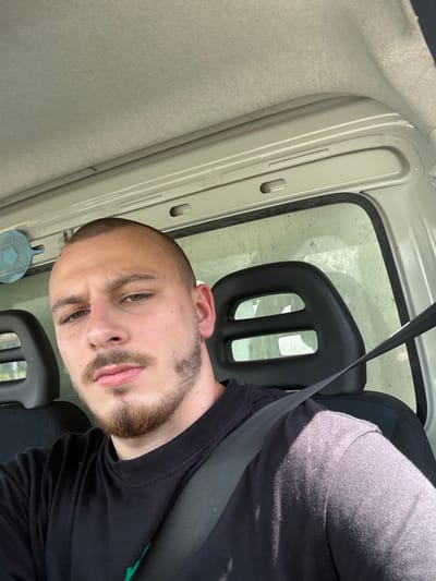 A close-up, vertical shot features a young adult male seated inside a vehicle in Kalitinovo, Bulgaria. The man, looking directly at the camera with a neutral expression, has short hair, a trimmed beard, and light-colored eyes. He wears a black t-shirt, and a black seatbelt crosses diagonally over his chest.
The setting is the interior of a car or van, indicated by the seatbelt, the design of the car seat's black headrest (which has two rectangular cut-outs), and the vehicle's light grey/beige roof lining and door frame components. A side window, visible behind the headrest, shows bright, sunlit conditions outside, though the glass appears speckled or dusty. Strong sunlight casts clear shadows, suggesting a sunny day. A small, blue, circular object with a grid pattern, possibly an air freshener or a decorative item, is attached near the bottom left of the man's head, close to the dashboard or door. There is no visible text in the image. The man appears to be taking a selfie or posing for a picture while stationary.