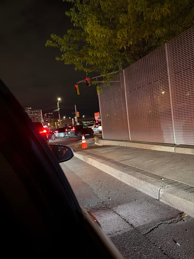 The image, taken at night from inside a vehicle, shows a scene of urban traffic stopped at a traffic light. The view is partially obscured by the interior of the car. Outside, cars are lined up in the road, illuminated by their brake lights. An orange traffic cone is visible near the side of the road. Above the road, a pedestrian bridge or overhead structure with a metal grid is visible. A tree with dense foliage is present on the left side of the frame. The location is inferred to be Fort Lee, New Jersey, based on the prompt. The road is alongside a curb and sidewalk, next to a wall or fence. Streetlights illuminate the area. Traffic lights show red.