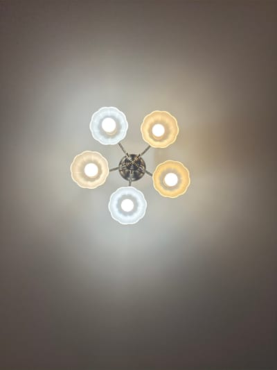 The image shows a ceiling-mounted chandelier with six lights arranged in a circular pattern. Three of the lights have frosted white glass shades, and the other three have frosted yellow glass shades. The chandelier is made of brushed metal. The ceiling is a plain gray color. The image is taken from below, looking directly up at the chandelier. There is no indication of the city of Rubtsovsk, Russia.