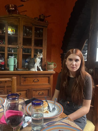 In Cañada de Madero, Mexico, a woman with long, reddish-brown hair is seated at a wooden table in front of a rustic wooden hutch. She is wearing a dark brown t-shirt and dark denim jeans. On the table, there is a plate with a partially eaten burger, a wine glass filled with red wine, and a tall glass of water. The hutch behind her is filled with glassware and decorative items, and a calico cat is perched on its surface. The room has an orange-toned wall and dark, textured stonework on the right side, suggesting a warm, home-like atmosphere.