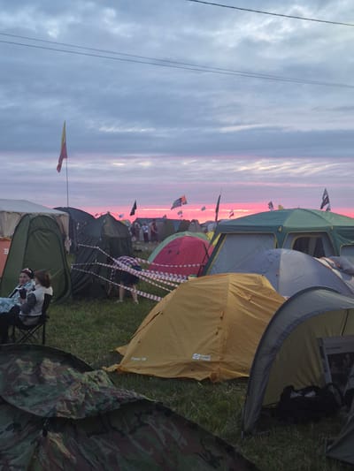 The image depicts a campsite at dusk, with a vibrant pink and orange sunset visible in the background. Numerous tents of various colors and sizes are set up on a grassy field. Some tents are green, yellow, red, and blue, with some featuring camouflage patterns. In the foreground, two women are sitting in chairs outside a green tent, with one of them looking towards the camera. Several other people are visible in the middle ground, some walking and others near tents. Red and white tape is strung between some of the tents, possibly to delineate pathways or sections of the campsite. Flags are flown on poles throughout the campsite, adding to the festive atmosphere. The overall impression is that of a busy festival or outdoor event.