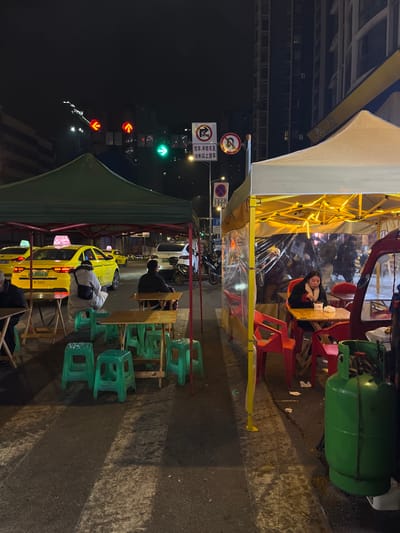 This nighttime image captures a vibrant street scene, likely an outdoor dining area or night market in an urban setting. The foreground features several temporary food stalls or restaurants operating under two distinct tents: a brightly lit yellow tent enclosed with plastic sheeting, and a darker green tent further down the street.
Under the yellow tent, a woman with dark hair is visible, leaning on a wooden table, looking towards the camera. She appears to be resting or waiting, surrounded by red plastic chairs. String lights illuminate the interior of this tent. Nearby, a large green gas cylinder stands on the pavement, suggesting cooking operations.
Under the green tent, several patrons are seated at wooden tables with green plastic chairs, some facing away from the camera. A yellow taxi cab is parked adjacent to this tent, its rear visible, bearing the Chinese characters "的士" (taxi) on its roof sign and a partially visible license plate starting with "G02". Other vehicles, including cars and motorcycles, are parked further along the street.
To the left of the scene, multiple traffic lights are visible: one green arrow indicating straight ahead, and three red arrow lights pointing left. Various road signs are also present, including a "No left turn" sign, a "No straight ahead" sign, and a circular "No stopping" sign. A blue rectangular sign clearly states "非机动车道" (Non-motorized vehicle lane), followed by "禁止非机动车通行" (No passage for non-motorized vehicles), and "20时至次日7时" (20:00 to 7:00 the next day), indicating time restrictions for non-motorized vehicles.
The ground appears damp, suggesting recent rain or evening moisture. In the background, illuminated buildings rise against the dark night sky. The overall atmosphere is bustling, characteristic of an evening eatery in an Asian city.