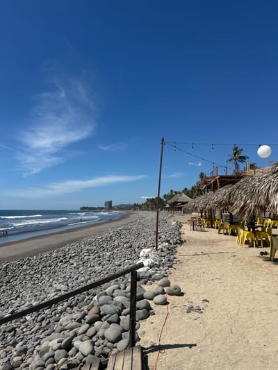 A rotated shot of a beach scene with a vibrant blue sky taking up a large portion. The foreground shows a rocky shoreline transitioning into a sandy beach. A structure with a thatched roof and yellow chairs sits atop the beach. A string of lights extends from the structure towards the foreground. In the distance, a building stands along the coastline. The ocean is visible in the middle ground with gentle waves breaking.