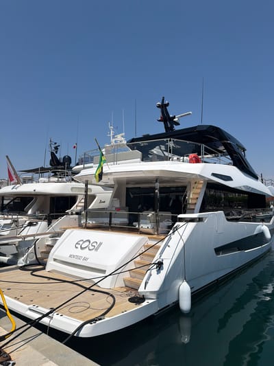 The image shows a large, modern white yacht docked in a marina. The yacht has a sleek, multi-level design with black accents and several windows. A wooden deck is visible at the stern, with ropes and mooring lines securing the vessel. There are life preservers on board. A Jamaican flag is flying from a mast on one of the yacht's decks. Text on the stern reads "EOSII MONTEGO BAY". Other yachts are visible in the background, and the water of the marina is calm and reflects the sky. The sky is clear and blue, suggesting a sunny day. The setting appears to be a harbor in Saint-Raphaël, France, given the location context. No people are clearly visible in the image. The scene is one of a quiet day at the harbor, with the focus on the impressive yacht.