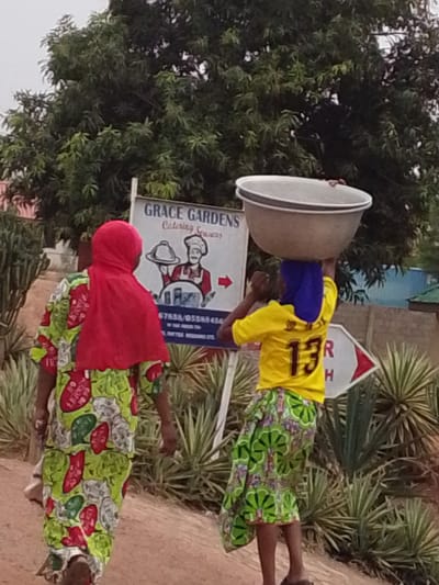 Two women walk on a dirt path in Dungu, Ghana. The woman on the left wears a red headscarf and a patterned green, red, and white dress. The woman on the right wears a blue headscarf, a yellow t-shirt with the number 13 on the back, and a green and flowered patterned skirt. She carries a large, silver bowl on her head. Behind the women, a sign reads "Grace Gardens Catering Services" with a chef illustration. An arrow points to the right on another sign. Green plants and trees are in the background.