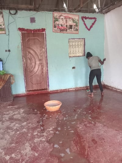 An interior view depicts an individual engaged in floor cleaning within a room in Manda-Shivanga ward, Kenya. The room features light blue/aqua painted walls, with a section of the right wall painted white, contrasting with the worn texture of the blue paint. A wooden door with a decorative circular panel is centrally located on the left wall, adorned with red tinsel garland above and along its frame. Exposed wiring and a light switch are visible near the door. The ceiling structure reveals exposed wooden beams and what appears to be corrugated iron roofing. Decorative elements on the walls include a large framed picture of a modern house with a red car, a smaller framed image below it, and red tinsel arranged in a triangular shape. The red/maroon concrete floor is extensively wet with foamy water, indicating ongoing scrubbing. An orange plastic basin, containing water, rests on the wet floor. To the far left, a portion of a television screen and a wooden cabinet are visible. An adult individual, observed from behind, is barefoot, wearing a light-colored t-shirt and dark trousers, and actively scrubbing the floor with a long-handled brush.