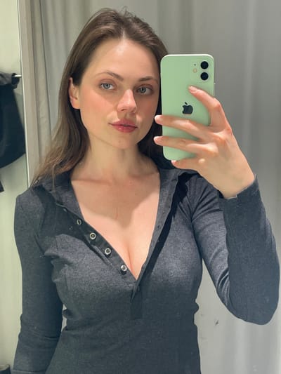 A young adult female with fair skin, medium-brown hair, and blue-green eyes is depicted in a mirror selfie. She wears a dark grey, long-sleeved, ribbed Henley-style top with a V-neckline, secured by five metallic snaps, two of which are open. A faint linear scar is present on her upper right chest. She holds a light green Apple iPhone with dual rear cameras in her right hand, oriented horizontally. The background features light-colored, vertically paneled walls, consistent with an indoor dressing room or changing area. A dark garment or object is partially visible hanging on the left. There is no visual evidence within the image to suggest a location in Pompei, Italy.