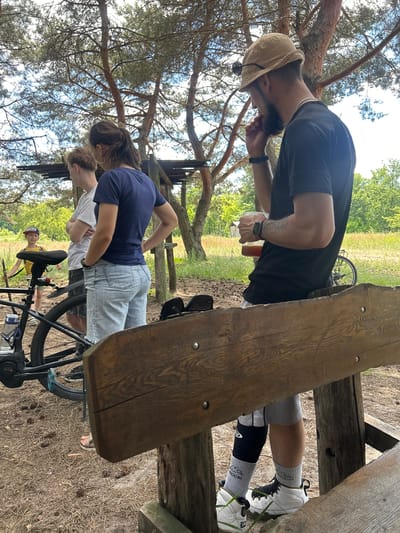 A group of people are gathered near a wooden structure and a bicycle in a wooded area. A man in a black t-shirt and a beige hat is holding a glass of amber liquid. To his left, a woman in a blue t-shirt and denim shorts stands with her hands on her hips. A young person in a white t-shirt is next to her. Further to the left, a young boy in a yellow shirt and a red baseball cap is holding a stick near a black bicycle. The ground is covered with pine needles and pine cones. In the background, trees and lush green foliage are visible. The overall setting suggests an outdoor recreational or picnic area. The location is specified as Chmyrivka, Ukraine.