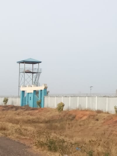 The image displays a security perimeter in Kerang, Nigeria, under a hazy, pale gray sky. A multi-level metal watchtower, topped with a blue, hip-roofed canopy and accessed by an external metal staircase, stands prominently on the left. Below the watchtower and integrated into a light gray concrete perimeter wall is a bright blue gate structure, featuring white crenellated coping. The entire length of the perimeter wall is topped with concertina wire. The foreground consists of arid, reddish-brown earth, sparsely covered with dry, light-brown vegetation and several columnar succulent plants. The edge of an unpaved road is visible in the lower-left portion of the frame. No individuals or active subjects are present.