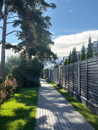 A paved walkway runs alongside a dark wooden fence, leading away from the viewer. Lush green grass lines the left side of the walkway, with ornamental bushes and large trees creating shade. The walkway is illuminated by dappled sunlight, casting geometric shadows. Two individuals are visible walking on the pathway in the distance, appearing as small figures. The fence, constructed of horizontal dark planks, encloses a property, and beyond it, modern residential buildings with green coniferous trees are visible. The sky above is a bright blue with scattered white clouds. The scene depicts a well-maintained residential area with a clear demarcation between public and private spaces.