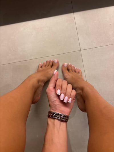 The scene depicts a top-down view of a person's lower body, specifically their bare feet, lower legs, and a single forearm and hand, situated on a tiled floor in Tumbak Bayuh, Indonesia. The individual's feet are positioned with toes pointing towards the top of the frame, showcasing light pink or nude painted toenails. The skin on the legs and feet appears tanned. The left forearm and hand are held forward, with the fingers curled slightly, displaying a French manicure with squared tips, featuring clear or light pink bases and white tips on all five visible fingernails. A metallic link-bracelet watch is worn on the left wrist. The environment consists of a light grey or beige tiled floor with visible grout lines. A shadow is cast from the upper left side of the frame.
