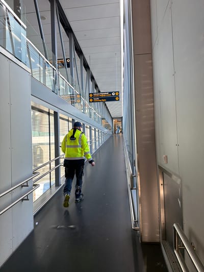 An indoor walkway at Oslo Airport Gardermoen, Norway, features a lone individual wearing a high-visibility yellow jacket with reflective stripes, black trousers, and a dark baseball cap. The person is walking away from the camera down the corridor, which has a dark floor and a silver-colored handrail on the left. Large glass windows line the left side of the walkway, offering a view of an exterior structure with metal supports and glass panels. The right side of the walkway is a plain gray wall. Overhead, signs are visible, one indicating "Ankomst / Bagagehall" (Arrivals / Baggage Hall) and "Transfer Connecting flights." The lighting suggests it is daytime, with natural light entering from the windows. The overall scene is clean and functional, characteristic of an airport environment.