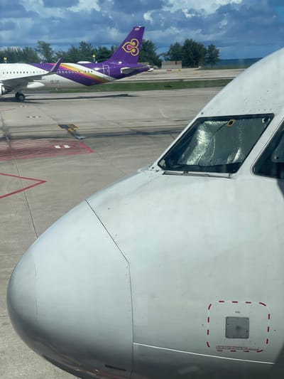 The image captures a close-up view of the nose and cockpit area of a white aircraft parked on an airport tarmac. The aircraft's fuselage is predominantly white, with visible rivets and panel lines. A sunshade is visible inside the cockpit windows, reflecting light. In the background, another aircraft with a distinctive purple and gold livery, identified as Thai Airways by its tail logo, is parked on the tarmac. The surrounding environment appears to be an airport with visible runway markings and distant trees, suggestive of a tropical location. The sky is partly cloudy. The location is identified as Mai Khao, Thailand.
