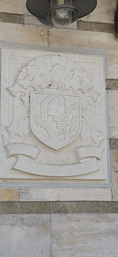 The image shows a stone relief of a coat of arms located in Cserszegtomaj, Hungary. The coat of arms is intricately carved with various symbols, including a shield with geometric shapes, what appears to be a bundle of wheat, and clusters of grapes. Flanking the shield are decorative elements that resemble foliage or possibly mythological figures. Below the shield, a banner inscription reads "CSERSZEGTOMAJ", with the years "1862" on the left and "2000" on the right, suggesting a historical significance or a commemoration. The relief is mounted on a light-colored stone wall, which is part of a building. A light fixture with a glass cover is visible at the top of the image, suggesting this is an outdoor installation. The surrounding stone blocks are textured and vary in color, with some darker green-hued stone interspersed. The overall impression is of a historical monument or building facade.