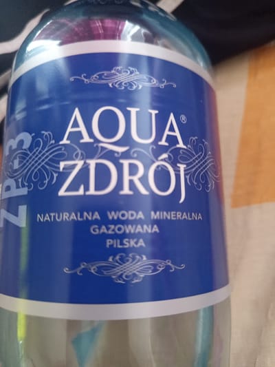 This is a close-up, eye-level, indoor photograph of a plastic bottle of "Aqua Zdrój" brand mineral water. The label is predominantly blue with white lettering and ornate white decorative elements. The text on the label reads "AQUA ZDRÓJ", and below that, "NATURALNA WODA MINERALNA GAZOWANA PILSKA". On the left side of the label, partially obscured, are the characters "ZP3". The bottle is positioned against a textured background that appears to be a piece of fabric, possibly a blanket or a bedspread, with areas of dark stripes and a light orange or beige color. The lighting is somewhat uneven, creating highlights and shadows on the bottle and the background. There is no indication of the city of Czersk, Poland, or any specific location within it in this image.