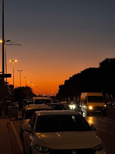 The image captures a street scene in Kartal, Turkey, during a twilight sunset. The sky is a gradient of deep blue at the top transitioning to a vibrant orange near the horizon, with a few faint stars visible. Lined up along the side of the road are several cars, some parked and others in traffic. The foreground features a white car, its headlights reflecting on the wet pavement, suggesting recent rain. Further down the street, a yellow van is illuminated by its headlights, also on the road. Streetlights cast a warm glow on the scene, enhancing the late evening atmosphere.

The activity depicted is everyday urban life, with vehicles navigating the street and people presumably going about their evening. The presence of multiple streetlights and the clear visibility of the cars suggest this is a well-lit urban environment. The silhouette of trees lines the right side of the road, contrasting with the bright orange sky. The overall mood is calm and serene, characterized by the beautiful sunset colors and the quiet hum of a city winding down for the night. There are no visible signs with text, but a "FIAT" logo is present on the white car in the foreground.