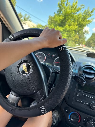 The image shows a person's hand on the steering wheel of a car. The steering wheel is covered with a black leather-like material and has decorative rhinestones. The car's dashboard is visible, including the speedometer, radio, and air conditioning controls. Outside the car, green trees and a blue sky can be seen, suggesting it is daytime and likely summer or spring. The image is taken from inside the car, looking forward. There is no indication of the specific location within Basarabeasca, Moldova.