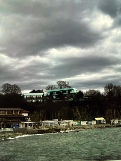 The image depicts a coastal scene under a dramatic, cloudy sky. In the foreground, the choppy, greenish-blue water of the sea is visible, with waves lapping against a rocky shoreline. Along the shore, there is a row of buildings and structures. To the left, a multi-story building with a reddish-brown roof and visible windows is present. Adjacent to it, a long, low-lying structure with a white pergola-like roof extends along the waterfront, featuring multiple enclosed spaces and open areas. Some of these structures appear to be shops or cafes, with signage and displays visible. Further along the coast, smaller, box-like structures with arched roofs are seen. The middle ground is dominated by a hillside covered in dark foliage and trees. On the hillside, several modern-looking buildings are situated. The most prominent of these is a multi-story building with a flat, light blue roof and white balconies. Another similar building, also with a light blue roof, is partially visible to the right. The background is filled with a heavily overcast sky, characterized by dark, textured clouds with patches of lighter grey and white, suggesting an impending storm or heavy cloud cover. The overall impression is one of a somewhat somber, overcast day at a seaside resort or coastal town. The location is not the city of Artik, Armenia.