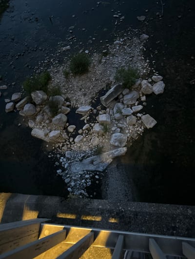 This image provides an aerial, high-angle view of a shallow riverbed in Anduze, France. The riverbed is characterized by a scattering of light-colored rocks and pebbles, with patches of dark water flowing between them. Several small green bushes are visible among the rocks. In the foreground, a concrete structure with metal railings suggests a walkway or stairs leading down towards the river. The lighting indicates it is dusk or night, with artificial lights casting yellow and white beams onto the concrete and railings. The scene is calm and appears to be a natural environment within the city.