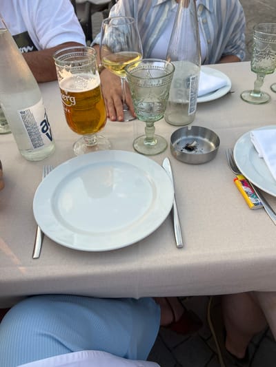 The image depicts a table set for a meal, likely outdoors given the natural lighting and casual attire. The scene is set on a beige tablecloth, with several elements suggesting a relaxed dining experience.
On the table, there are multiple drinking glasses, some containing pale yellow liquid, possibly white wine or beer, with one glass holding a frothy beer. There are also two white plates, one with a white napkin neatly folded on top, and a fork resting on it, alongside a colorful disposable lighter. A metal ashtray filled with cigarette butts sits near the center of the table. A clear glass bottle of mineral water, with visible condensation, is also present.
Two individuals are partially visible in the frame. One person, wearing a blue and white striped shirt, has their arm resting on the table. Another person, in a white t-shirt, is also visible. Further down the table, parts of legs are seen, with one person wearing blue clothing and the other wearing brown shoes, suggesting they are seated. The ground visible outside the table appears to be paved with dark stones, hinting at an outdoor patio or garden setting.
The lighting suggests it is daytime, possibly late afternoon or early evening, as the light appears soft and warm. The presence of an ashtray indicates that smoking is permitted in this area. The overall impression is one of a casual meal or gathering. Text on the mineral water bottle includes brand names and Serbian or a similar Slavic language text, potentially related to mineral content and origin.