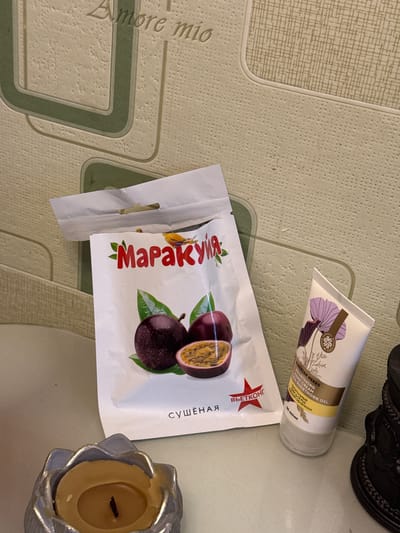 A flat-lay image shows several items on a surface. The top right corner contains a black, ornate object. Below it and angled towards the upper right is a white tube with beige and purple floral designs, labeled with the text "SIBERIAN HERBS NOURISHING HAND CREAM SEA-BUCKTHORN OIL". To the left and slightly lower is a white package with a picture of passion fruit and the word "Маракуйя" in red Cyrillic script. Beside this packet to the right sits a silver candle holder with a lit candle. The surface has a light green patterned design, and "Amore mio" is partially visible in the bottom left. There is no location specified in the city of Ketoshi, Russia, nor any people or interactions visible in the image.