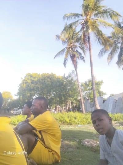 Three men are sitting on a grassy field in Nungwi, Tanzania, located at coordinates -5.726, 39.301. They are all wearing casual clothing. Two of the men are wearing yellow shirts, and one is wearing a white shirt. There are palm trees in the background. The man in the foreground is looking directly at the camera. He is wearing a white shirt and has a light complexion. His arms are resting on his legs. The man in the middle is wearing a yellow shirt and has a darker complexion. He is looking away from the camera, to the left. The man in the background is wearing a yellow shirt and has a darker complexion. He is also looking away from the camera, to the left. There is a partial view of another man in the background. He is wearing a black shirt and is sitting on the grass. The camera is facing the three men. There is a "Galaxy A05s" label in the lower left corner of the photo.